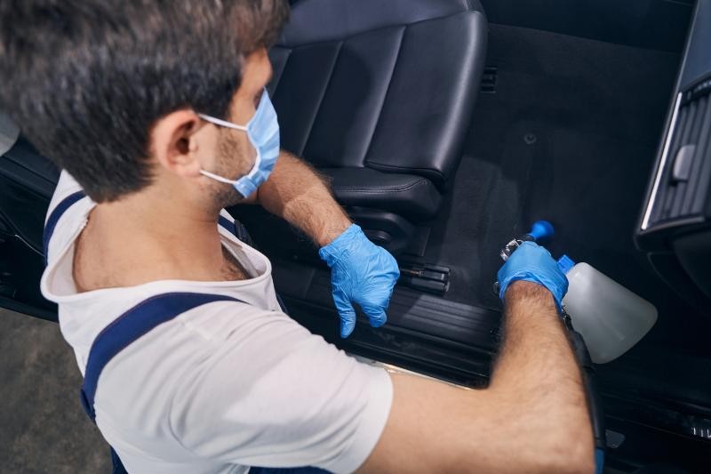 Professional detailer in gloves sanitizing vehicle interior seats with spray during mobile car detailing
